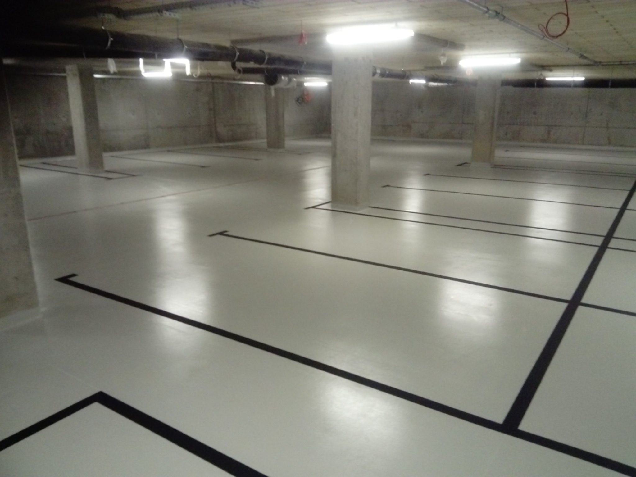 Jasnoszara posadzka z czarnymi liniami wyznaczającymi miejsca parkingowe w garażu podziemnym, widoczne betonowe kolumny i instalacje pod sufitem.