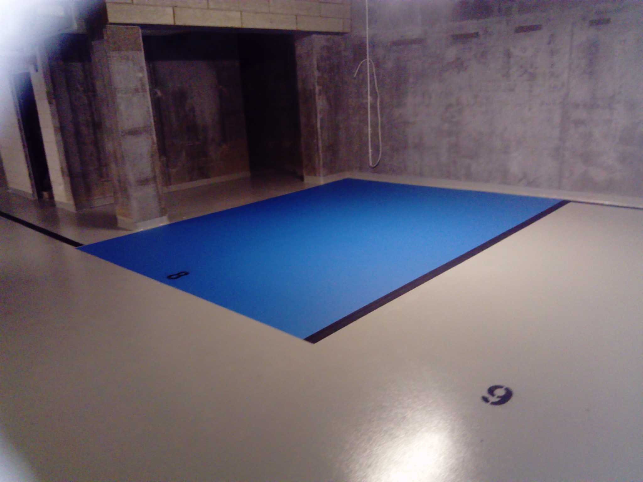 Wykończona posadzka przemysłowa z żywicy, z wyznaczonym niebieskim obszarem i numerami 8 i 9, w surowym wnętrzu z betonowymi filarami i ścianami.