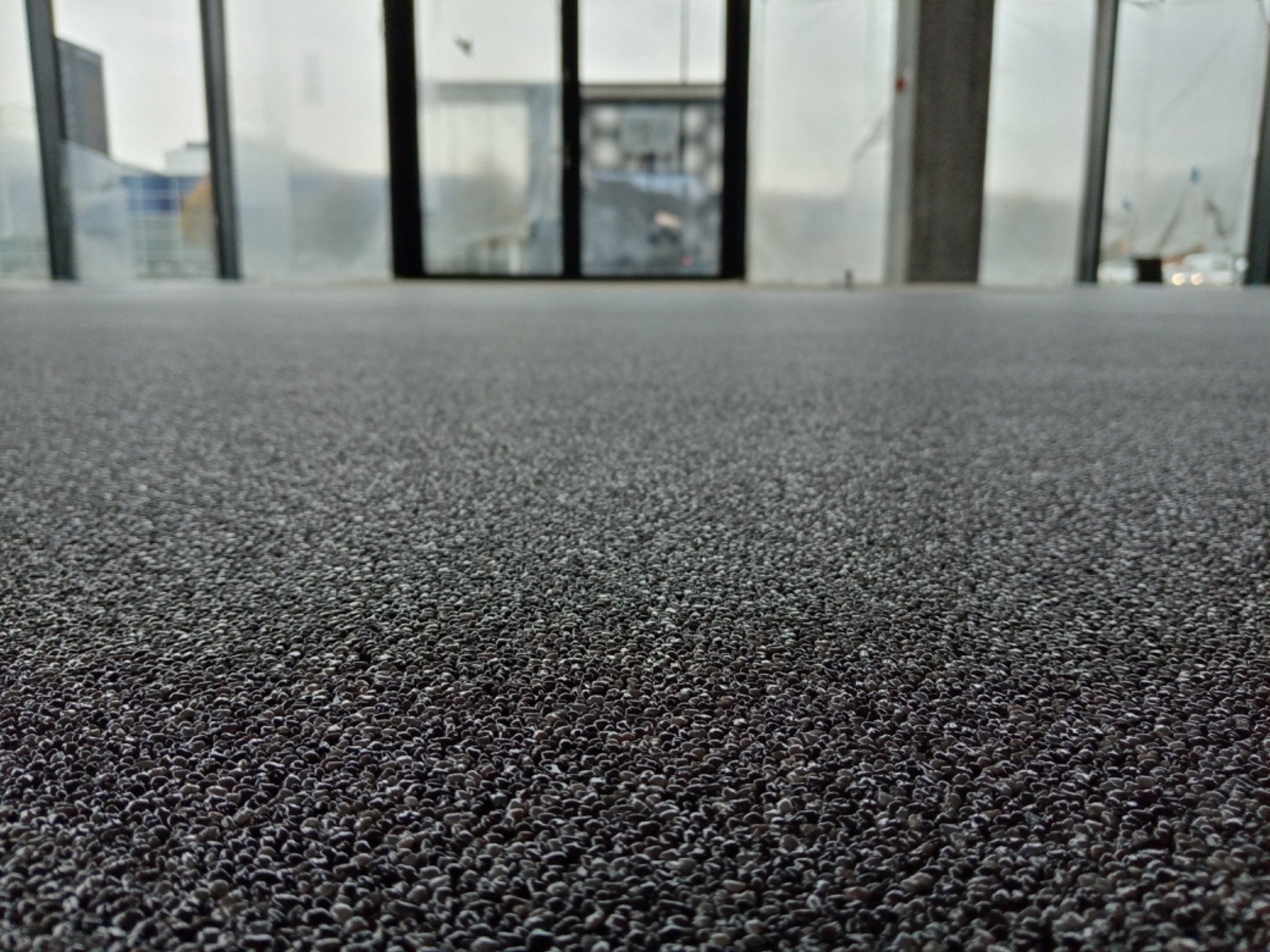 Szara, drobnoziarnista podłoga w pomieszczeniu z dużymi oknami i drzwiami balkonowymi w tle.