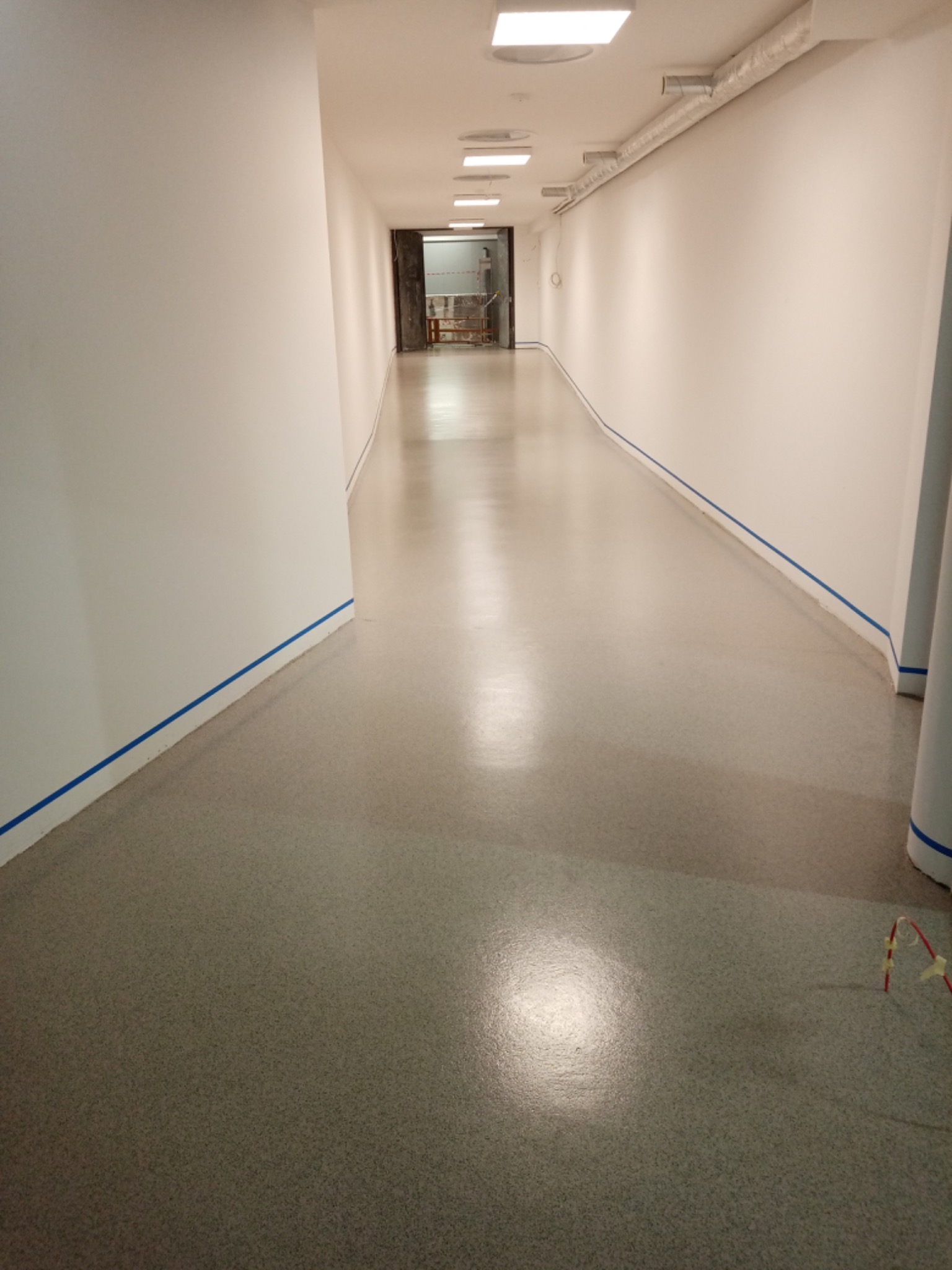 Długi korytarz z szarą, gładką posadzką, zabezpieczoną niebieską taśmą przy ścianach, prowadzący do otwartego pomieszczenia z widocznymi elementami konstrukcyjnymi.