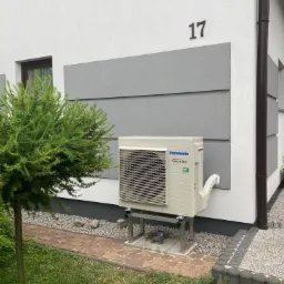 Zewnętrzna jednostka klimatyzacji Panasonic Inverter R32 zamontowana na metalowym stelażu przy ścianie domu z numerem 17, obok ozdobnego iglaka.