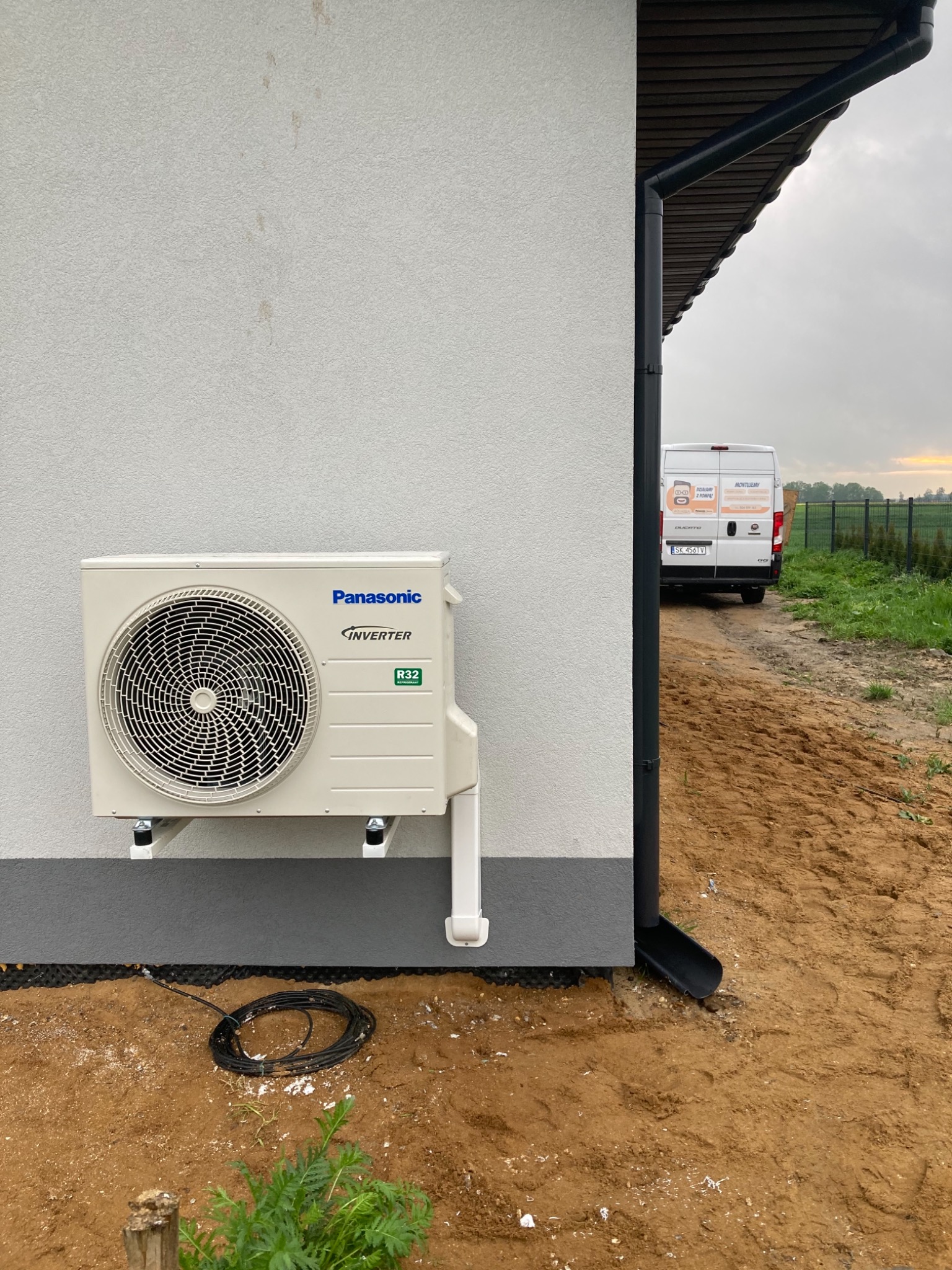 Zewnętrzna jednostka klimatyzacyjna Panasonic z widocznym napisem Inverter i symbolem R32, zamontowana na elewacji budynku z widocznym samochodem dostawczym w tle.