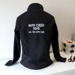 Czarna kurtka softshell z nadrukowanym białym logo 'AUTO CZĘŚCI TOYO' i numerem telefonu, leżąca na białej komodzie. Widoczny tył kurtki.