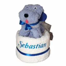 Ręcznikowy szary piesek z niebieską kokardą i imieniem Sebastian wyhaftowanym na białym ręczniku, całość zapakowana w celofan.