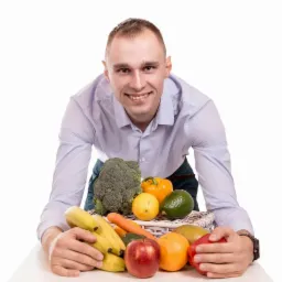 Uśmiechnięty mężczyzna w niebieskiej koszuli prezentuje koszyk pełen różnorodnych owoców i warzyw na białym tle.