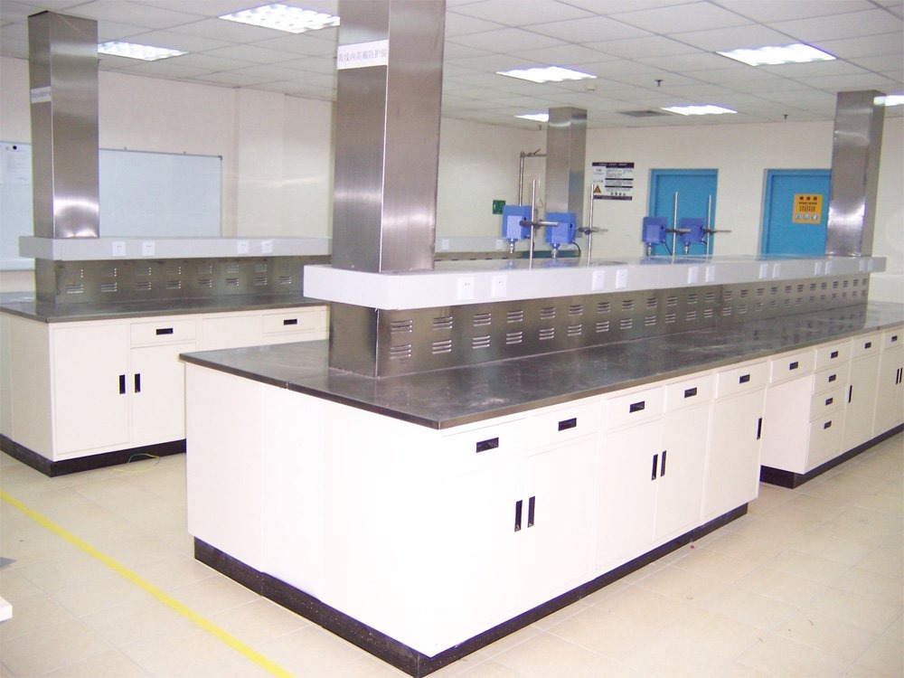 Wnętrze laboratorium z białymi szafkami i metalowymi blatami, widoczne stalowe kominy wentylacyjne, żółta linia na podłodze wyznacza strefę bezpieczeństwa.