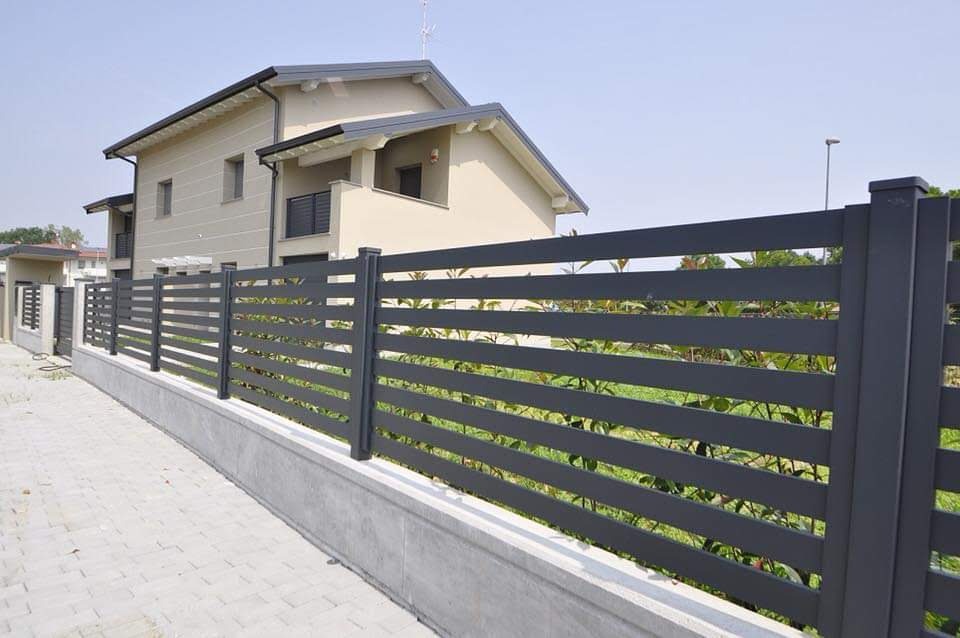 Nowoczesne, poziome ogrodzenie panelowe w kolorze grafitowym, zamontowane na niskim betonowym murku, na tle domu jednorodzinnego z jasną elewacją i szarym dachem.