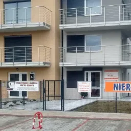 Budynek biurowy z balkonami i szyldem firmy projektującej wnętrza i ogrody, widoczne ogrodzenie z metalowej siatki i zaparkowane samochody.