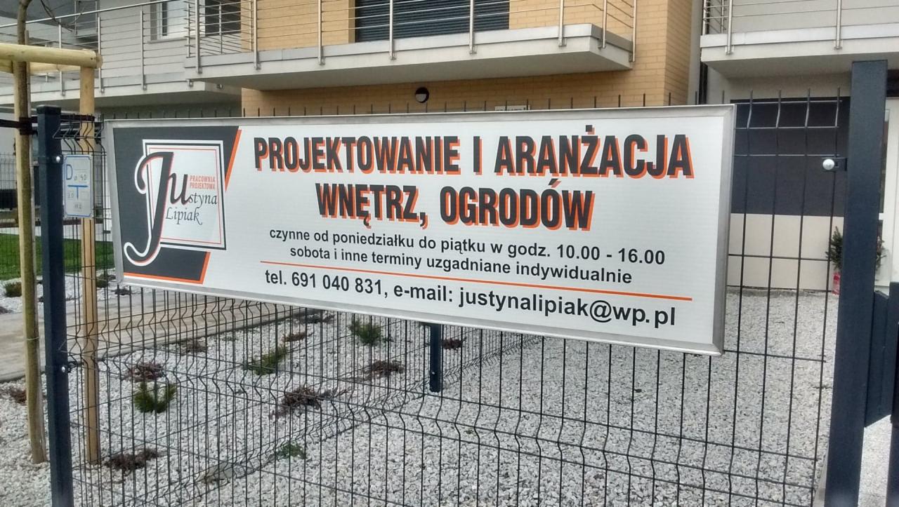 Szyld reklamowy firmy projektującej wnętrza i ogrody, umieszczony na ogrodzeniu z siatki na tle budynku.