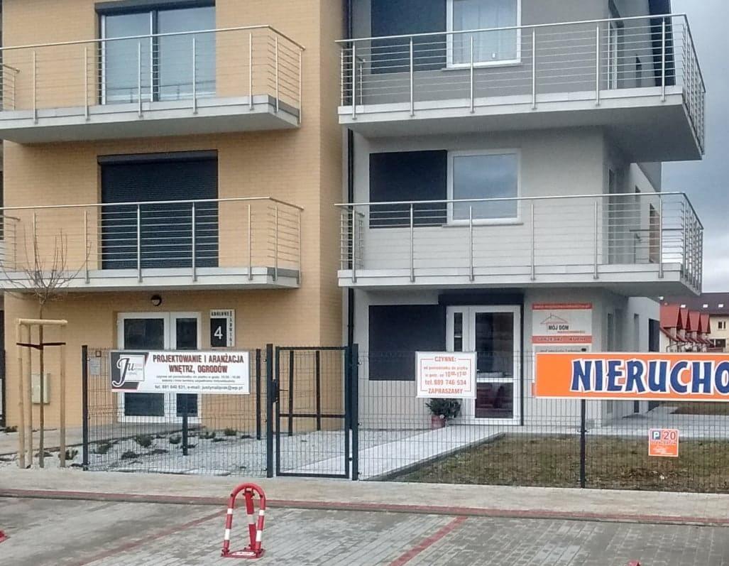 Budynek biurowy z balkonami i szyldem firmy projektującej wnętrza i ogrody, widoczne ogrodzenie z metalowej siatki i zaparkowane samochody.