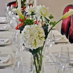 Uroczysty stół z białym obrusem, nakryty białą zastawą, kryształowymi kieliszkami i wazonem z białymi kwiatami (hortensje, lilie, dalie) oraz płonącymi świecami w srebrnych świecznikach.
