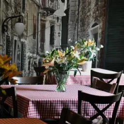 Ustawienie stołu z czerwono-białym obrusem w kratę, wazonem z liliami i tłem imitującym kamienną uliczkę, przygotowane do cateringu.