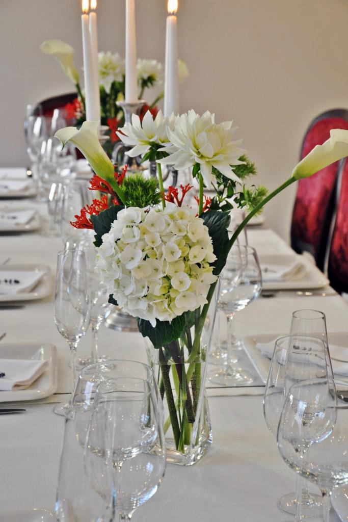Uroczysty stół z białym obrusem, nakryty białą zastawą, kryształowymi kieliszkami i wazonem z białymi kwiatami (hortensje, lilie, dalie) oraz płonącymi świecami w srebrnych świecznikach.