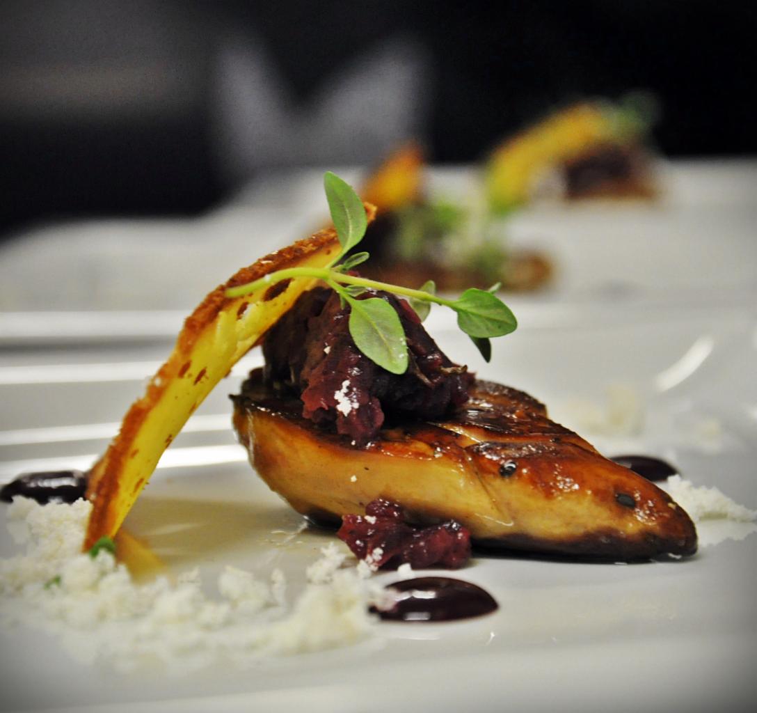 Elegancko podane danie z foie gras z konfiturą z czerwonej cebuli, grzanką i dekoracją z ziół na białym talerzu, z widocznymi elementami dekoracyjnymi z sera i sosem balsamicznym.