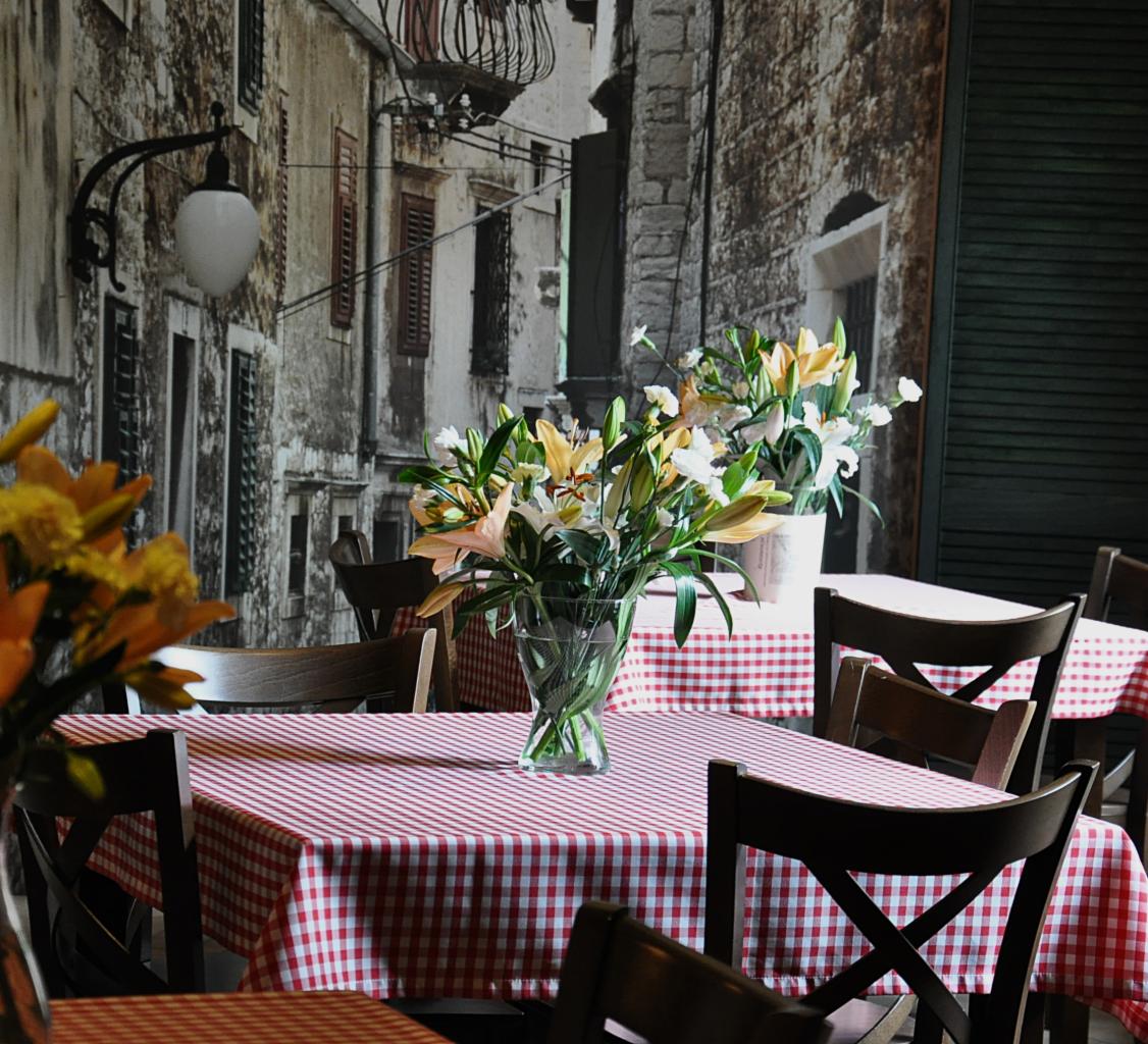 Ustawienie stołu z czerwono-białym obrusem w kratę, wazonem z liliami i tłem imitującym kamienną uliczkę, przygotowane do cateringu.