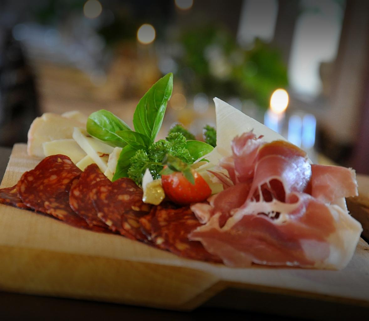 Deska serów i wędlin z chorizo, szynką parmeńską, serem oraz dekoracją z bazylii i pomidorka koktajlowego, podświetlona świecą w tle.