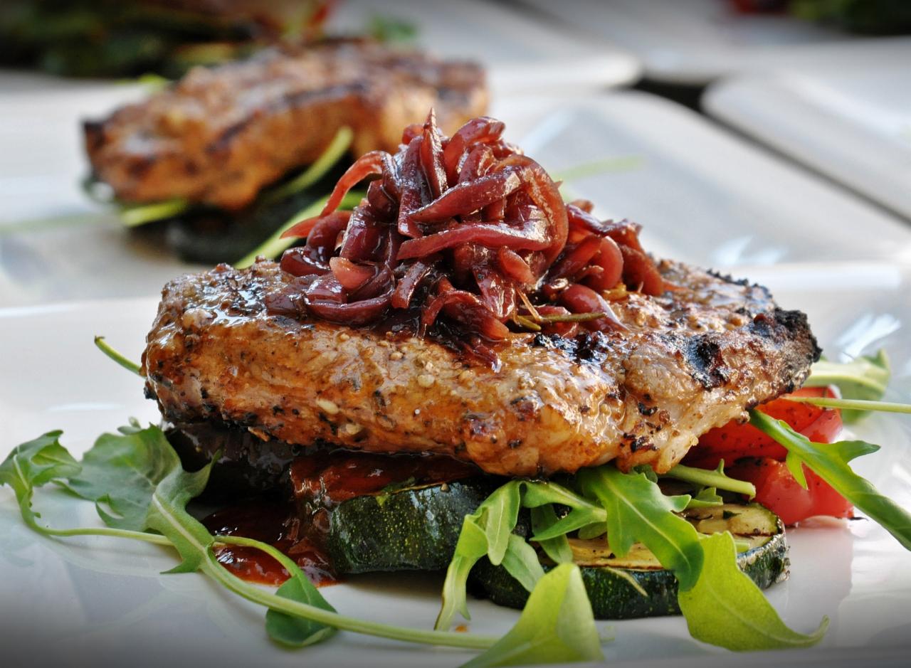 Apetycznie wyglądający stek z grilla, udekorowany karmelizowaną czerwoną cebulą i świeżą rukolą, podany na grillowanej cukinii, na białym talerzu.
