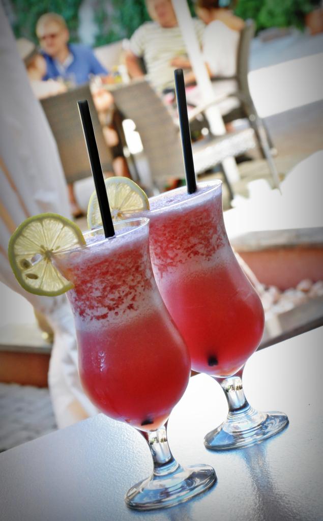 Dwa koktajle z czerwonego sorbetu w wysokich szklankach, ozdobione plasterkami limonki i czarnymi słomkami, na stole w plenerowej restauracji.