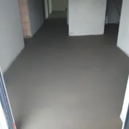 Świeżo wylana, gładka posadzka w surowym stanie w pomieszczeniu z widocznymi ścianami w trakcie wykończenia i surowym murem z cegieł w tle.