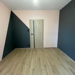 Wnętrze pokoju po odświeżeniu: ściany pomalowane w geometryczne wzory w odcieniach różu i granatu, jasna drewniana podłoga z paneli, nowoczesne drzwi z przeszkleniem.