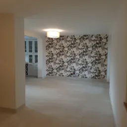 Wykończone wnętrze z szarą podłogą z płytek i ścianą pokrytą tapetą w motywy kwiatowe i ptasie, widoczna przeszklona szafka w tle.