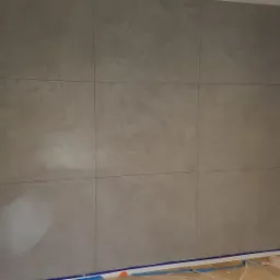 Ściana pokryta szarą, dekoracyjną gładzią, podzielona na prostokątne panele, zabezpieczona taśmą malarską przy podłodze i futrynie drzwi.