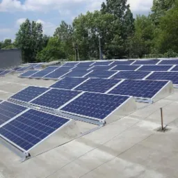 Rząd paneli słonecznych na płaskim dachu budynku przemysłowego, widoczne elementy montażowe i uziemienie, w tle drzewa.