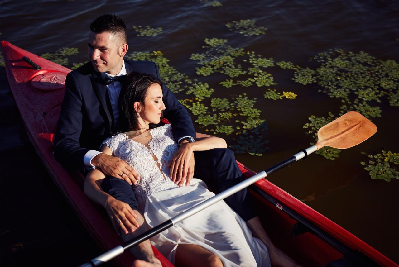 Elegancko ubrana para relaksuje się w czerwonym kajaku na tle ciemnej wody z pływającymi roślinami.