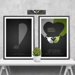 Dwa plakaty oprawione w czarne ramy, stojące na białym stoliku. Lewy plakat przedstawia szare serce na czarnym tle, prawy - zielono-szare ręce tworzące serce, również na czarnym tle. Oba plakaty...