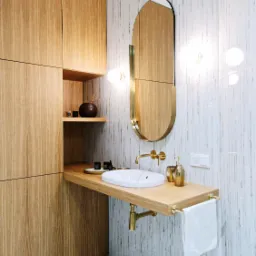Nowoczesna łazienka z drewnianymi szafkami, owalnym lustrem w złotej ramie i białą umywalką na drewnianym blacie z złotą armaturą, uzupełniona o biały ręcznik i dekoracyjne akcesoria.