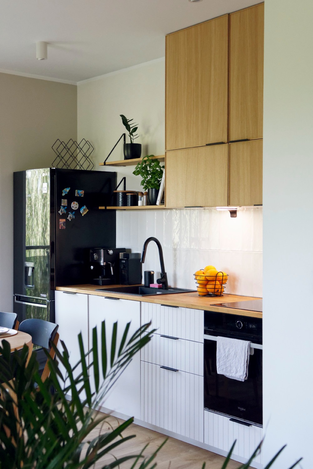 Nowoczesna kuchnia z drewnianymi szafkami wiszącymi, białymi szafkami dolnymi z pionowymi żłobieniami, czarnym zlewem i baterią, czarną lodówką z magnesami, oraz roślinami doniczkowymi na półkach.