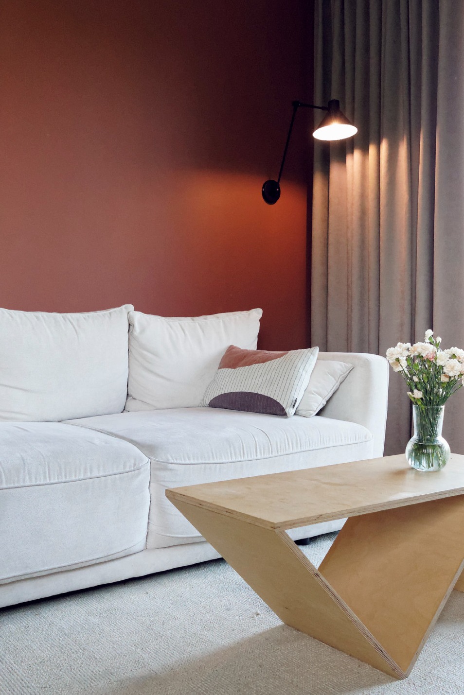 Jasny salon z białą sofą, geometrycznym stolikiem kawowym z jasnego drewna i ścianą w kolorze terakoty, oświetloną czarną lampą ścienną.