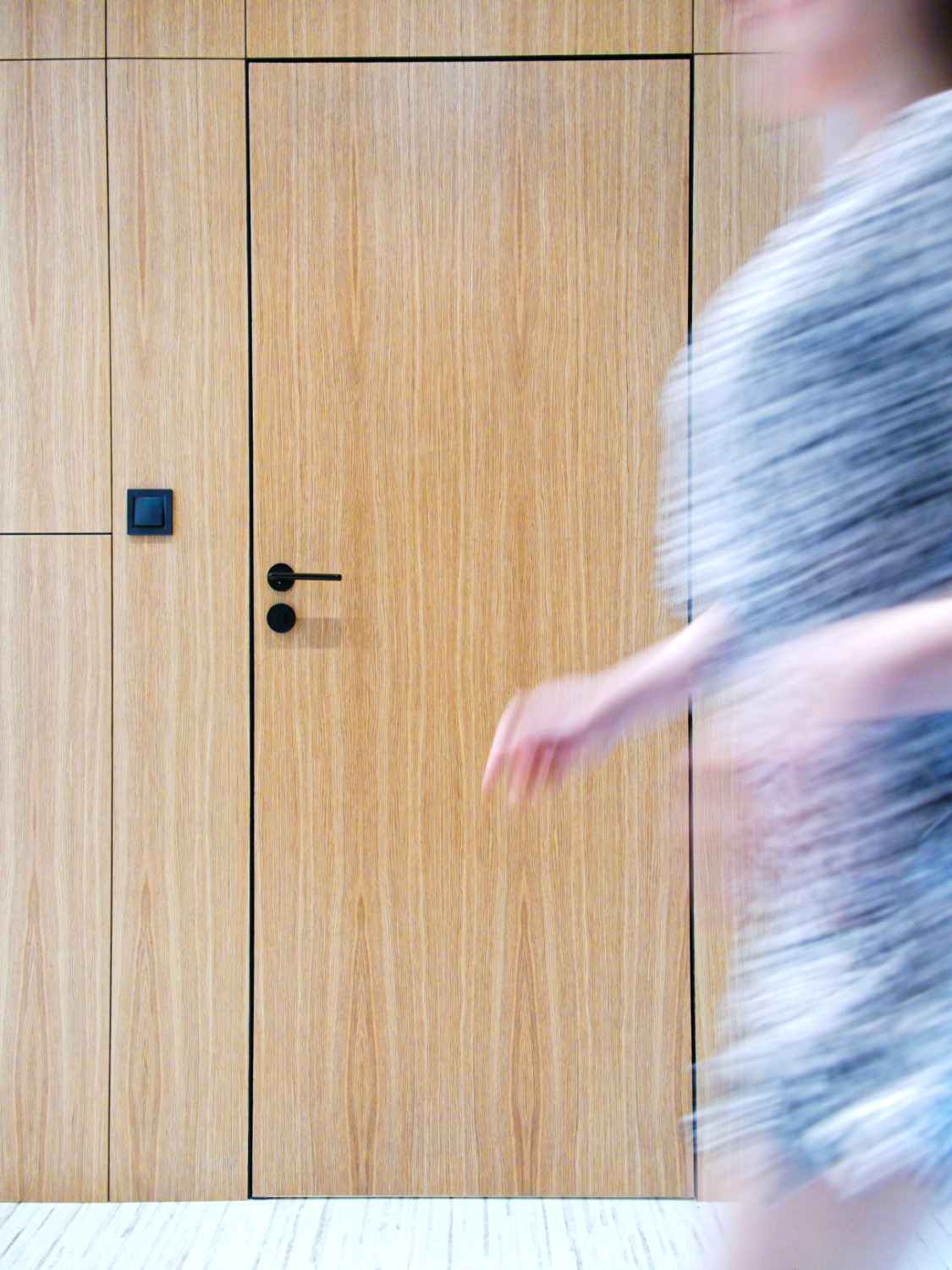 Minimalistyczna zabudowa ścienna z ukrytymi drzwiami z jasnego drewna, czarny włącznik i klamka, rozmyta sylwetka osoby w ruchu po prawej stronie.