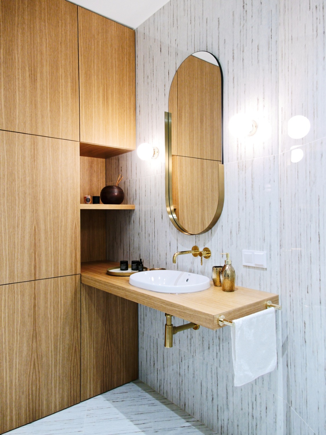 Nowoczesna łazienka z drewnianymi szafkami, owalnym lustrem w złotej ramie i białą umywalką na drewnianym blacie z złotą armaturą, uzupełniona o biały ręcznik i dekoracyjne akcesoria.