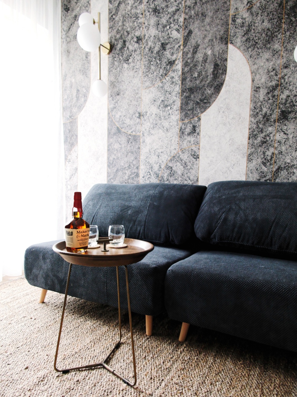 Nowoczesny salon z ciemnoniebieską sofą, stolikiem kawowym z butelką Maker's Mark i dwoma szklankami, na tle ściany z geometrycznym wzorem w odcieniach szarości i bieli oraz lampą kulową.