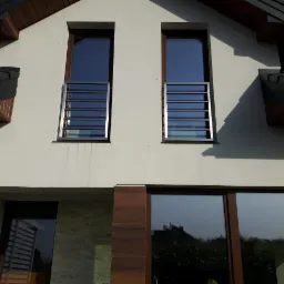 Nowoczesna elewacja domu z dwoma oknami na piętrze zabezpieczonymi poziomymi, metalowymi barierkami ochronnymi w kolorze srebrnym.