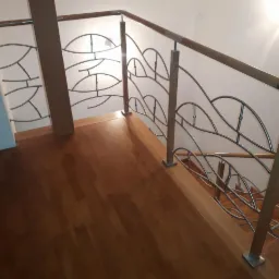 Nowoczesne schody z drewnianymi stopniami i balustradą z metalowymi elementami w kształcie stylizowanych liści, zamontowaną na drewnianych słupkach.