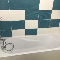 Biała wanna z chromowaną armaturą i słuchawką prysznicową na tle ściany wyłożonej biało-turkusowymi płytkami w szachownicę oraz drabinkowym grzejnikiem.