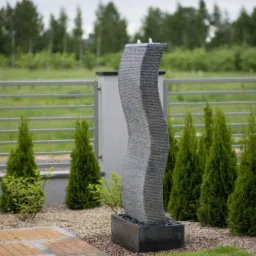 Nowoczesna fontanna ogrodowa z szarego kamienia o falistym kształcie, otoczona tujami i żwirem, z widoczną metalową siatką ogrodzeniową w tle.