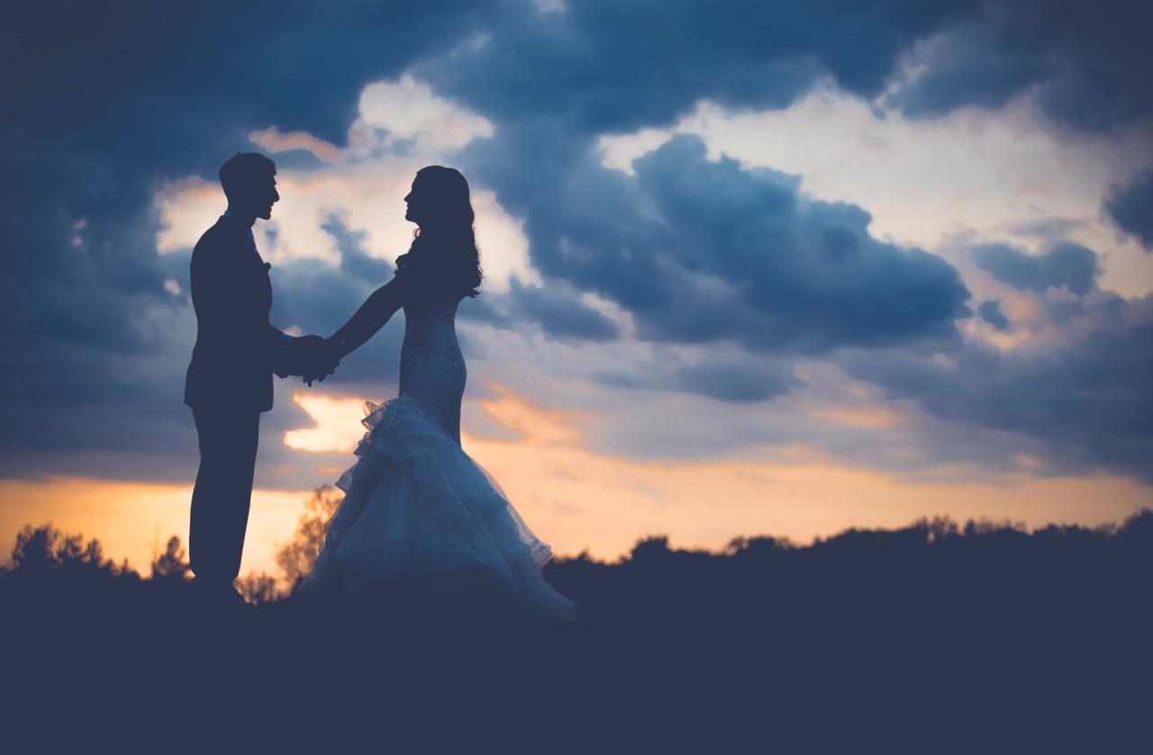 Silhouettes of a bride in a ruffled gown and groom holding hands at sunset, with dramatic cloudy sky.