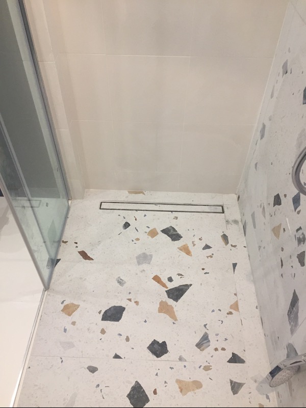 Biała łazienka z brodzikiem bezprogowym wykończona płytkami lastryko, widoczny odpływ liniowy i szklana kabina prysznicowa.