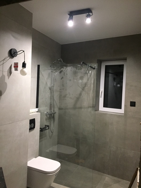 Nowoczesna łazienka z szarą glazurą, kabiną prysznicową typu walk-in z przezroczystą szybą, wiszącą toaletą, oświetleniem punktowym na suficie i kinkietem z żarówką Edison.