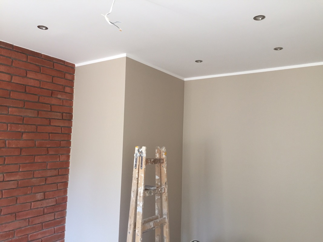 Ściana z cegły klinkierowej, pomalowane ściany w odcieniach beżu, biały sufit z oświetleniem punktowym i drewniana drabina malarska w rogu pomieszczenia.