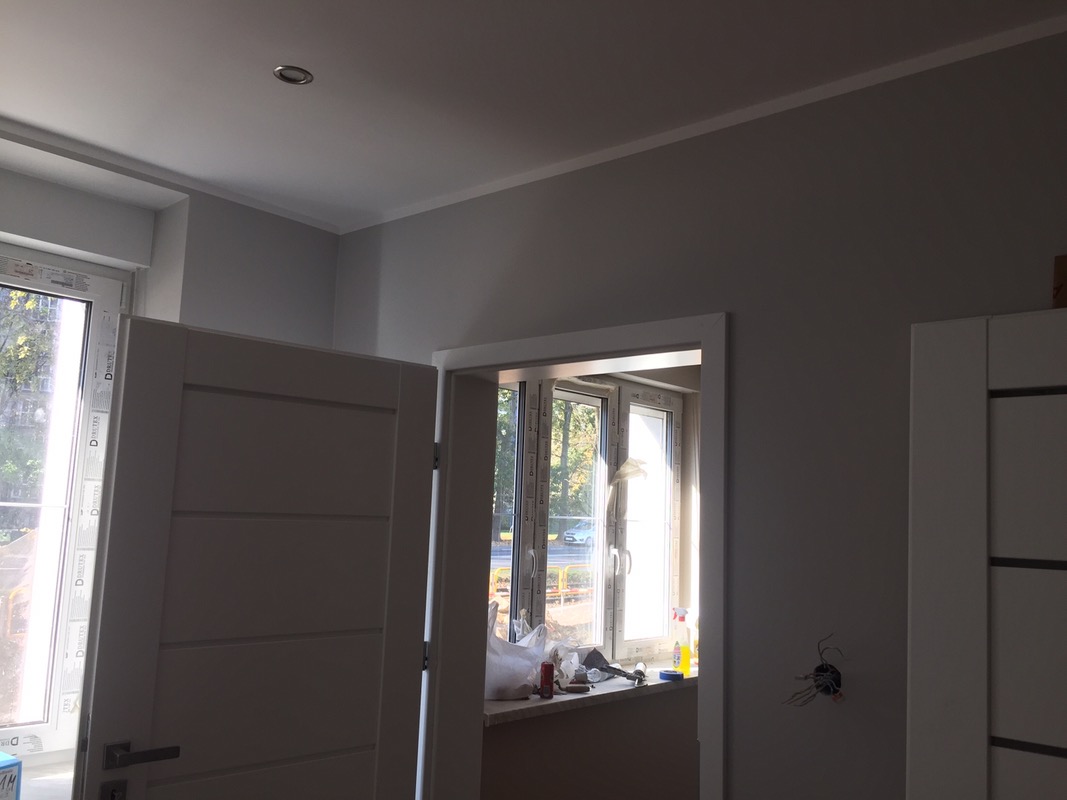 Szare wnętrze w trakcie wykończenia: widoczne drzwi wewnętrzne z poziomymi frezami, okno z parapetem, na którym leżą narzędzia i materiały budowlane, oraz wystające kable ze ściany.