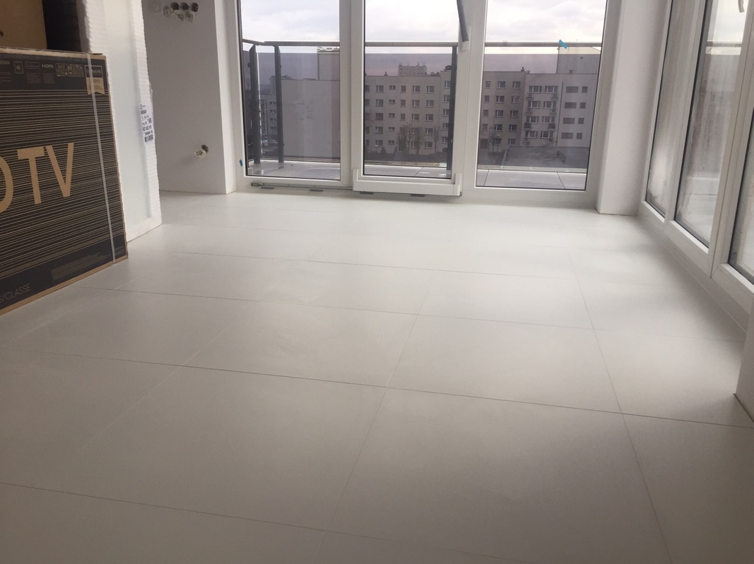 Jasnoszara, świeżo położona podłoga z dużych płytek w nowym mieszkaniu, widok na balkon i budynki za oknem, karton po telewizorze oparty o ścianę.