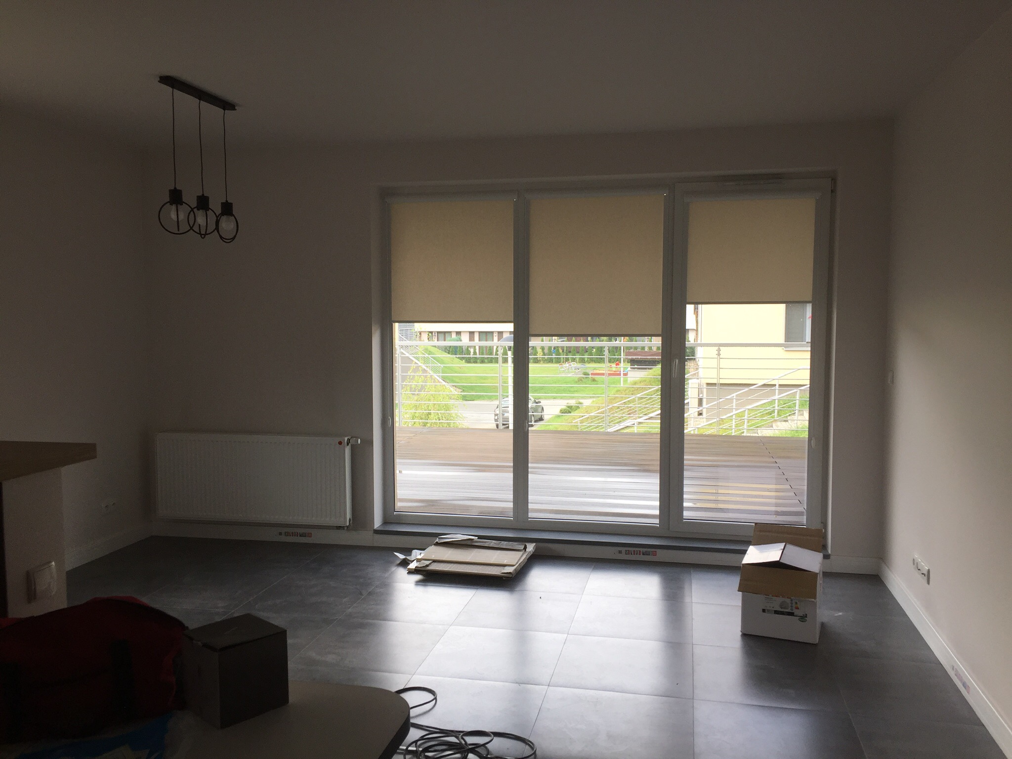 Pusty pokój z szarymi płytkami na podłodze, dużym oknem balkonowym z beżowymi roletami, wiszącą lampą z trzema żarówkami i grzejnikiem na ścianie. Na podłodze leżą kartony, co sugeruje niedawne...
