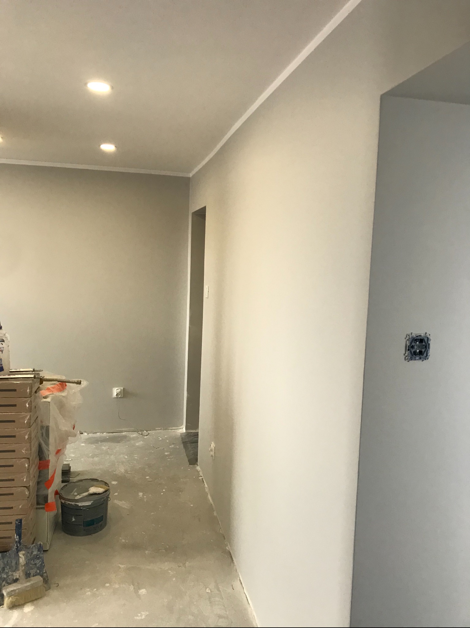 Wnętrze w trakcie malowania na szaro, widoczne puszki z farbą, wałki malarskie, zabezpieczone podłogi i surowe gniazdko elektryczne.