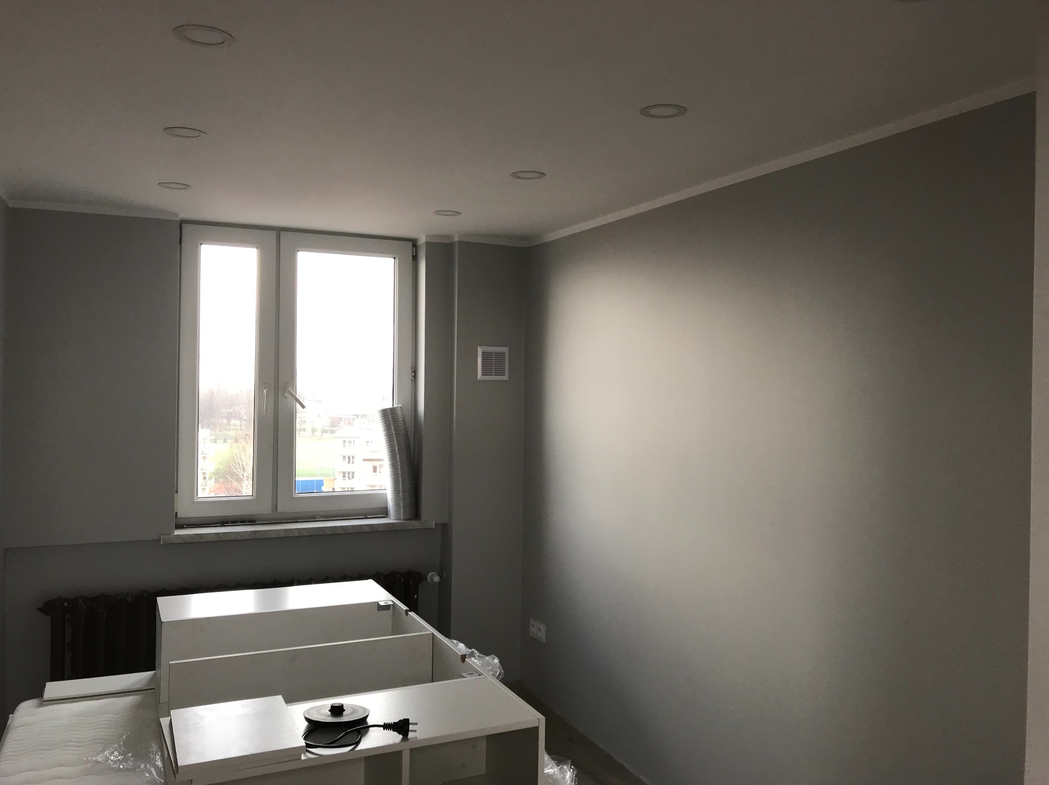 Szary pokój przed montażem mebli: widoczne białe szafki w folii, grzejnik pod oknem i sufit z wpuszczonymi lampami.