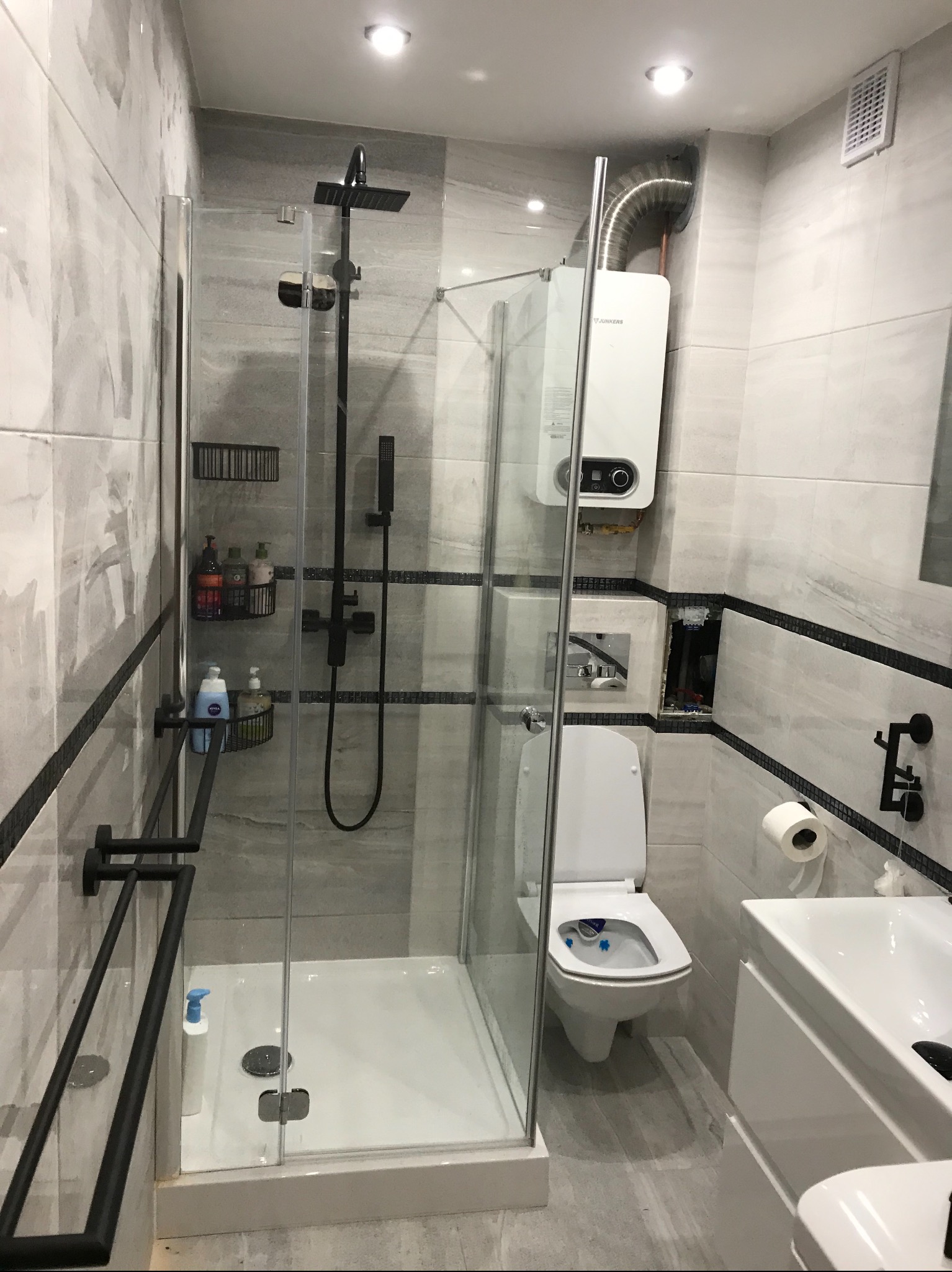 Nowoczesna łazienka z kabiną prysznicową typu walk-in, wiszącą toaletą z niebieskimi kostkami odświeżającymi, grzejnikiem Junkers i szarymi płytkami imitującymi marmur z czarnym paskiem dekoracyjnym.