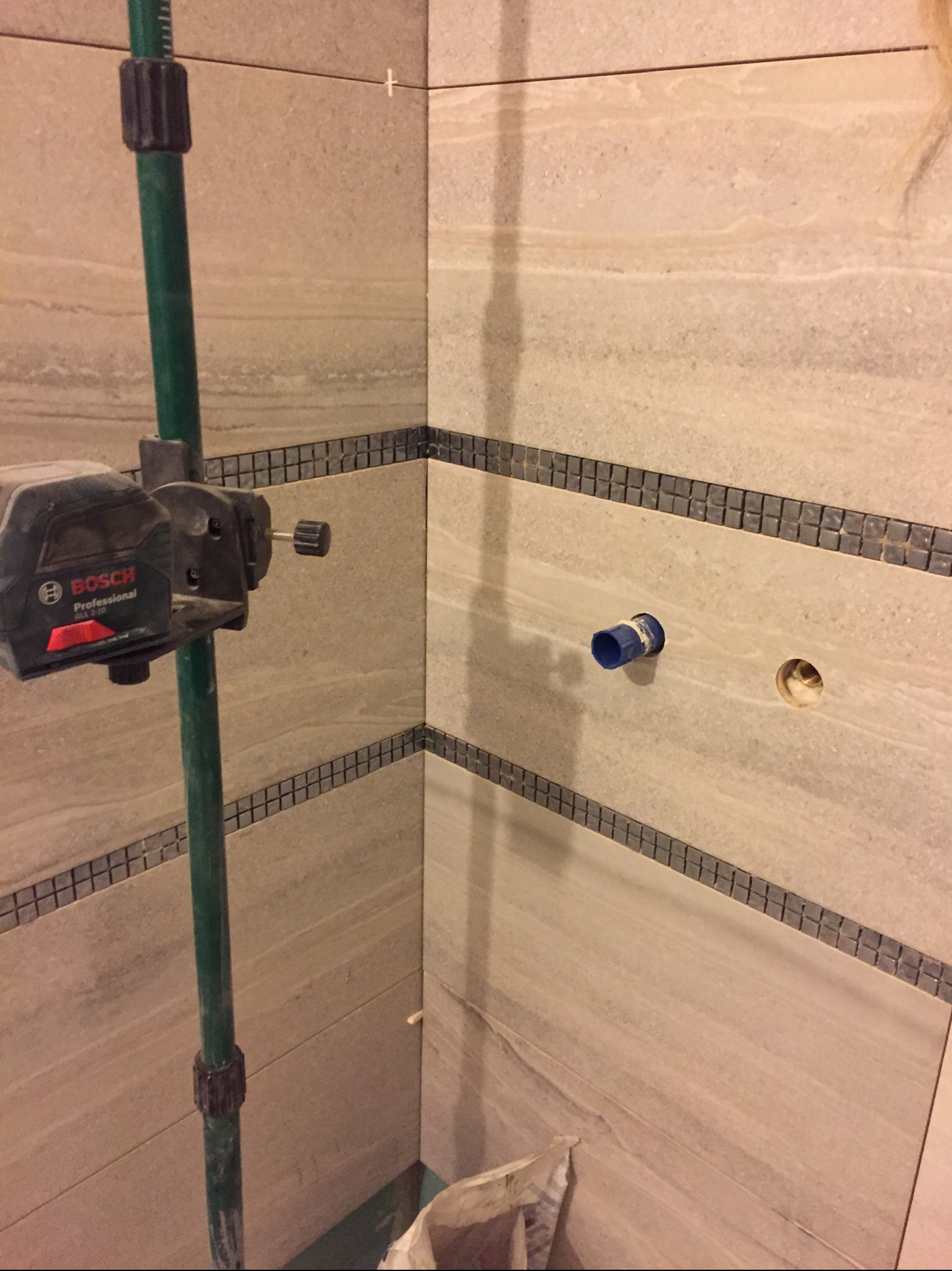 Fragment ściany łazienki z beżowymi płytkami imitującymi kamień, z dekoracyjnym pasem mozaiki, z widocznym otworem na rurę i zamontowanym niebieskim króćcem, poziomica laserowa Bosch na statywie...